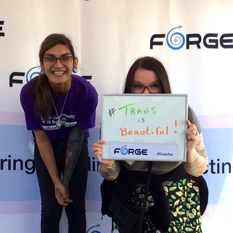 two smiling people holding a handwritten sign saying 'Trans is beautiful!' in front of a FORGE logo backdrop