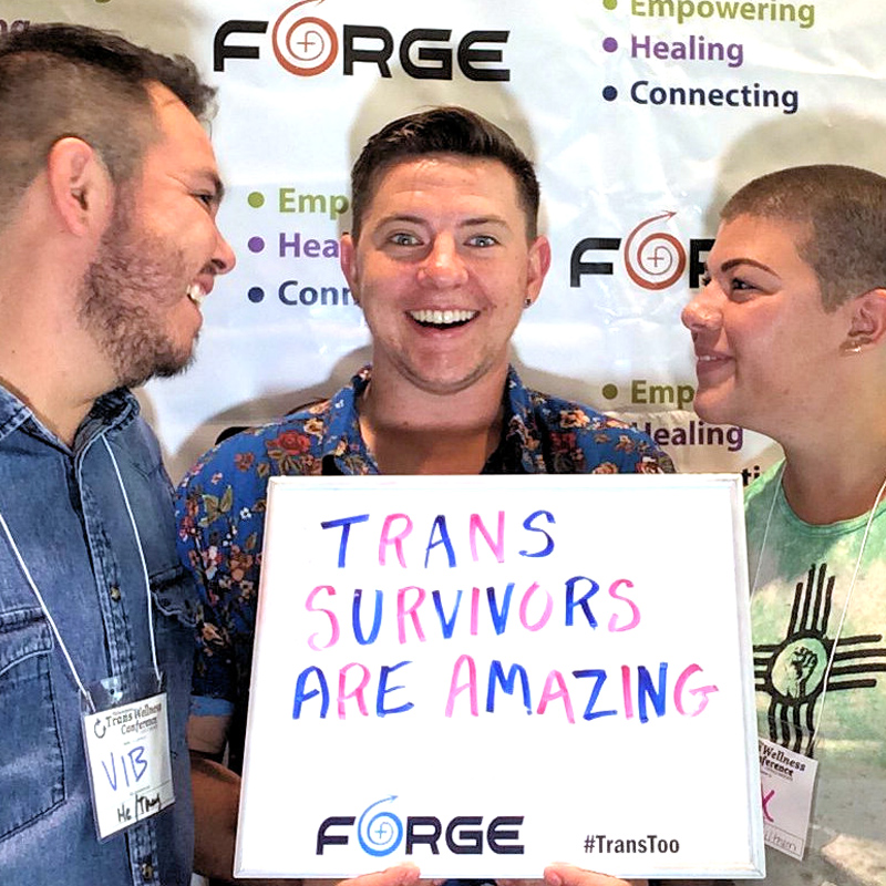 three laughing people holding a sign saying 'Trans Survivors are Amazing!' in front of a FORGE logo backdrop