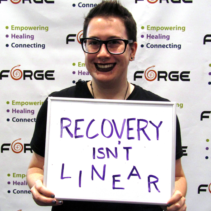 one smiling person holding a sign saying 'Recovery Isn't Linear' in front of a FORGE logo backdrop