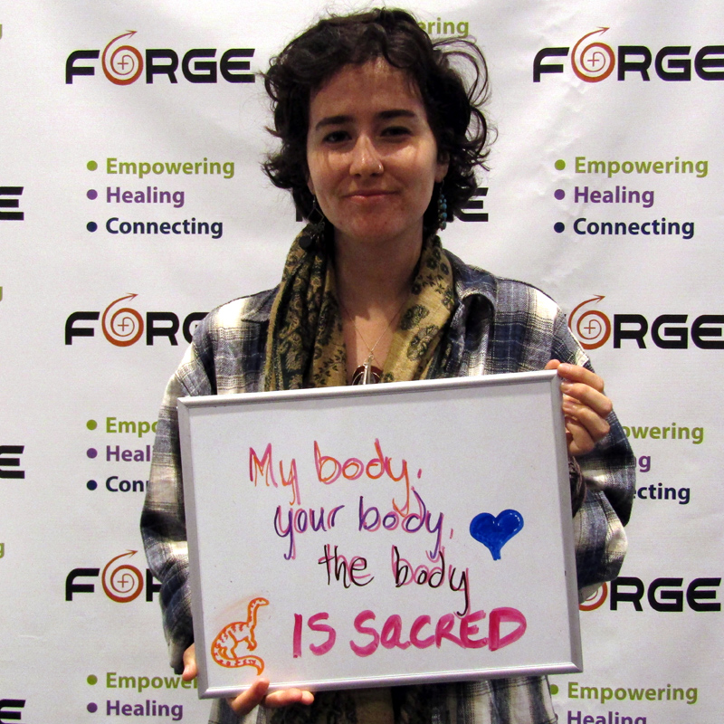 one smiling person holding a sign saying 'My body, your body, the body is sacred' in front of a FORGE logo backdrop