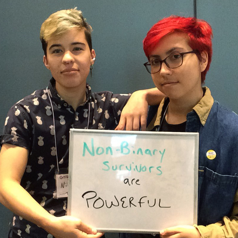 Two people holding a sign saying 'Non-Binary Survivors are Powerful!"